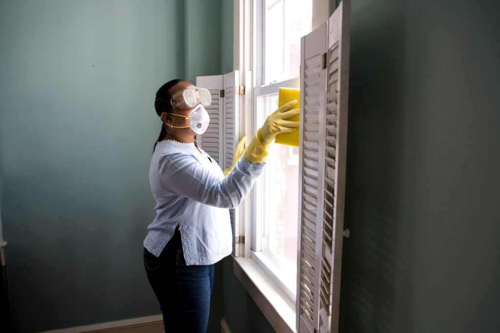 A person wearing a protective face mask cleaning windows inside a house